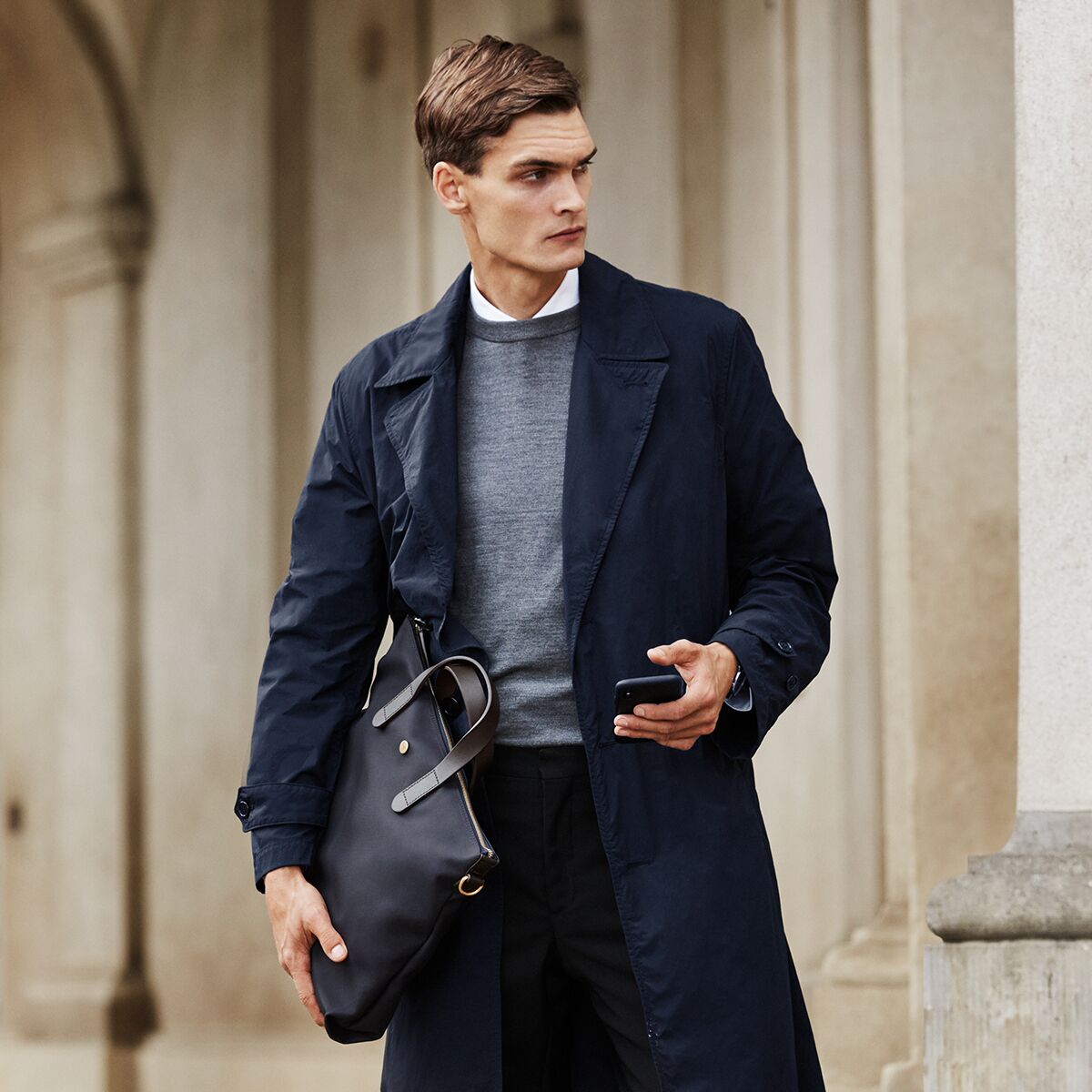 Ein eleganter Mann steht vor einer Steinarchitektur und trägt eine dunkelblaue MISMO Laptop-Tasche »BRIEF« mit braunen Lederdetails in der linken Hand. Er trägt einen dunkelblauen Mantel, grauen Pullover und weiße Hemdkragen, wirkt gepflegt und geschäftlich.