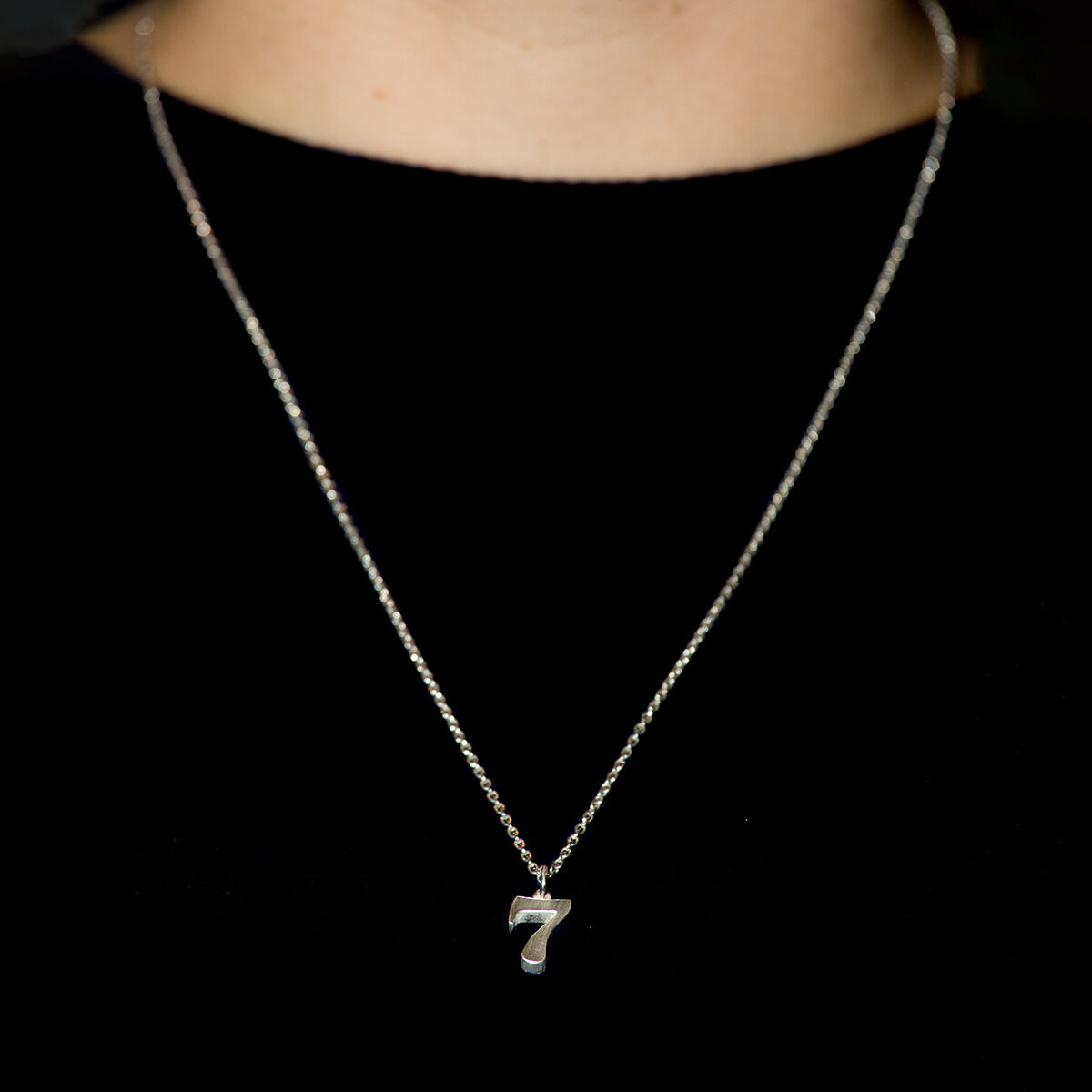 Nahaufnahme eines silbernen Zahlenanhängers „7“ aus massivem 925er Silber an zarter Kette, getragen an einem Hals vor schwarzem Hintergrund. Der Anhänger stammt aus der ZEIT-Sonderedition von Anke Gralfs und wird individuell handgefertigt.