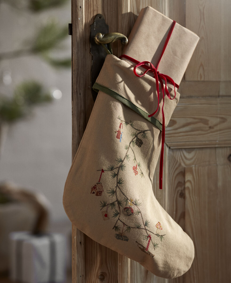 Beiger Weihnachtsstrumpf mit besticktem Tannenzweig und weihnachtlichen Motiven hängt an einer Holztür, darin ein in braunes Geschenkpapier eingepacktes, rotes Band umwickeltes Geschenk. Im Hintergrund ein unscharfer Weihnachtsbaum und ein weiteres Geschenkpaket.