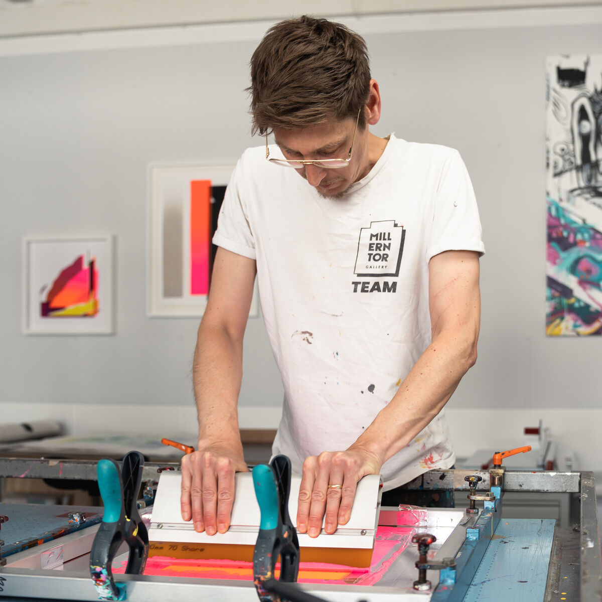 Im Vordergrund steht ein Mann in weißem, leicht verschmutztem T-Shirt mit Druck 'Millentor TEAM' an einem Siebdruck-Tisch, konzentriert beim Arbeiten. Im Hintergrund hängen gerahmte, farbenfrohe Bilder an einer hellen Atelierwand.