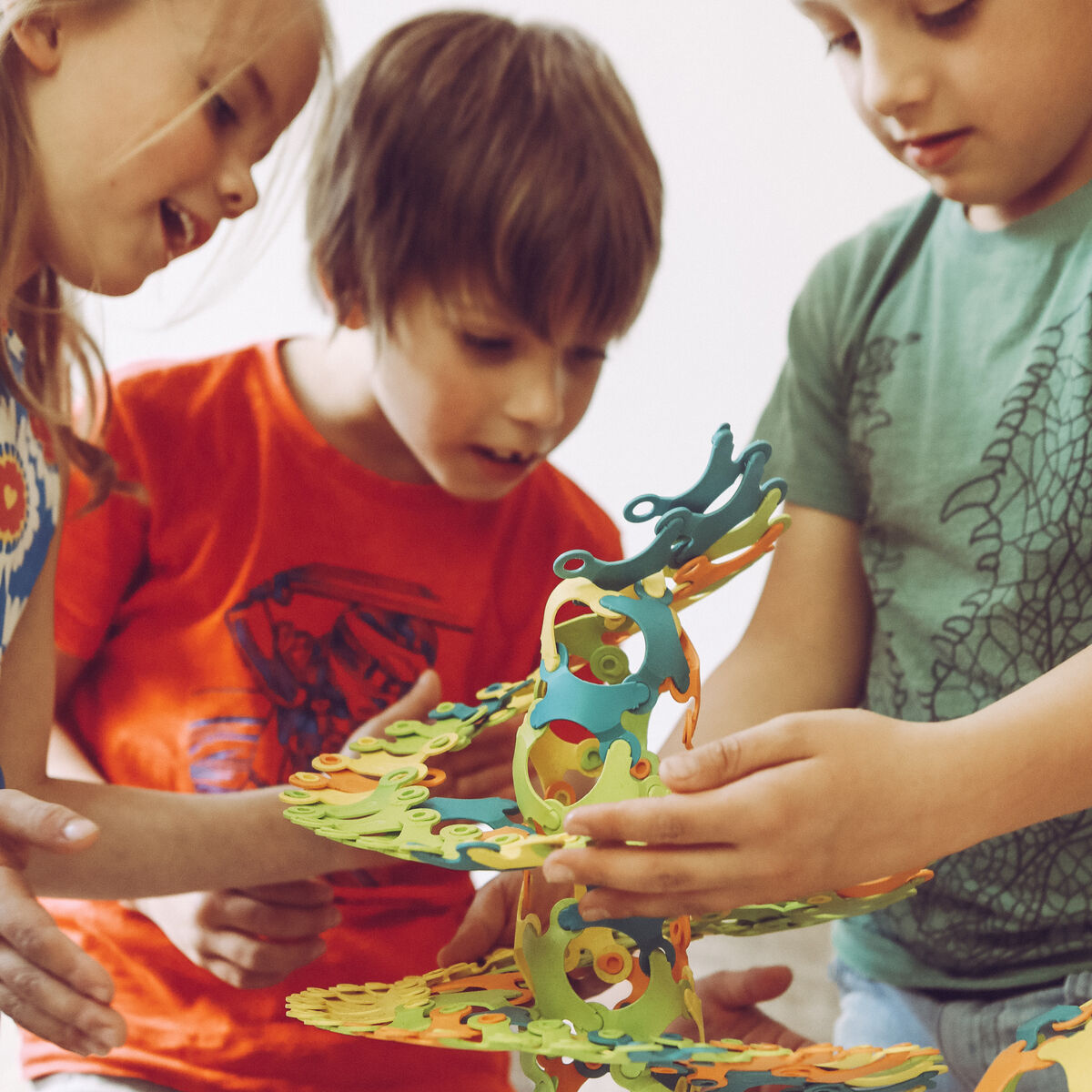 Drei Kinder spielen mit dem Konstruktionsspielzeug »Binabo« und bauen eine spiralförmige Struktur aus bunten Teilen in grün, orange, blau und gelb.