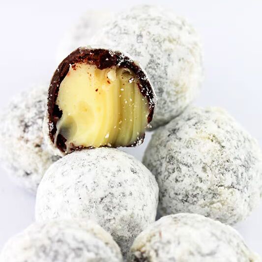 Mehrere Trüffelpralinen mit Champagnerfüllung liegen nebeneinander, eine Praline ist halbiert und zeigt eine helle Cremefüllung. Die Pralinen sind mit Puderzucker bestäubt und werden in einer Holzbox angeboten.