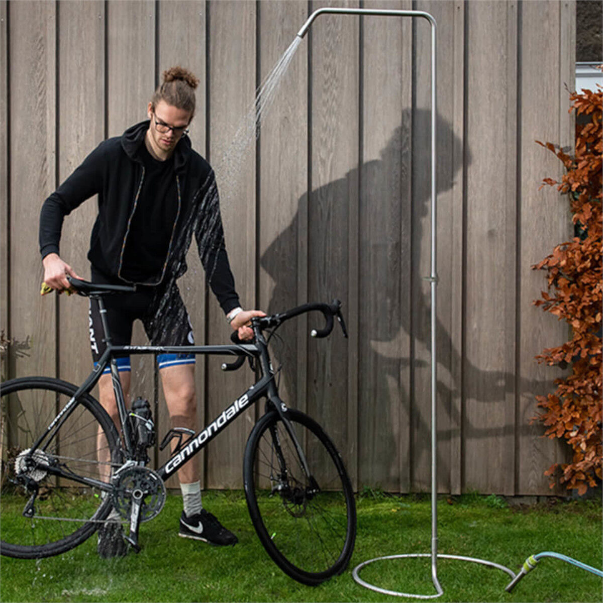 Ein Mann mit schwarzer Sportkleidung wäscht sein Fahrrad unter einer Gartendusche. Die Gartendusche ist an einem silbernen Rohr befestigt und steht auf einer grünen Rasenfläche neben einer Holzlattenwand. Im Hintergrund ist ein orangefarbener Strauch zu sehen.