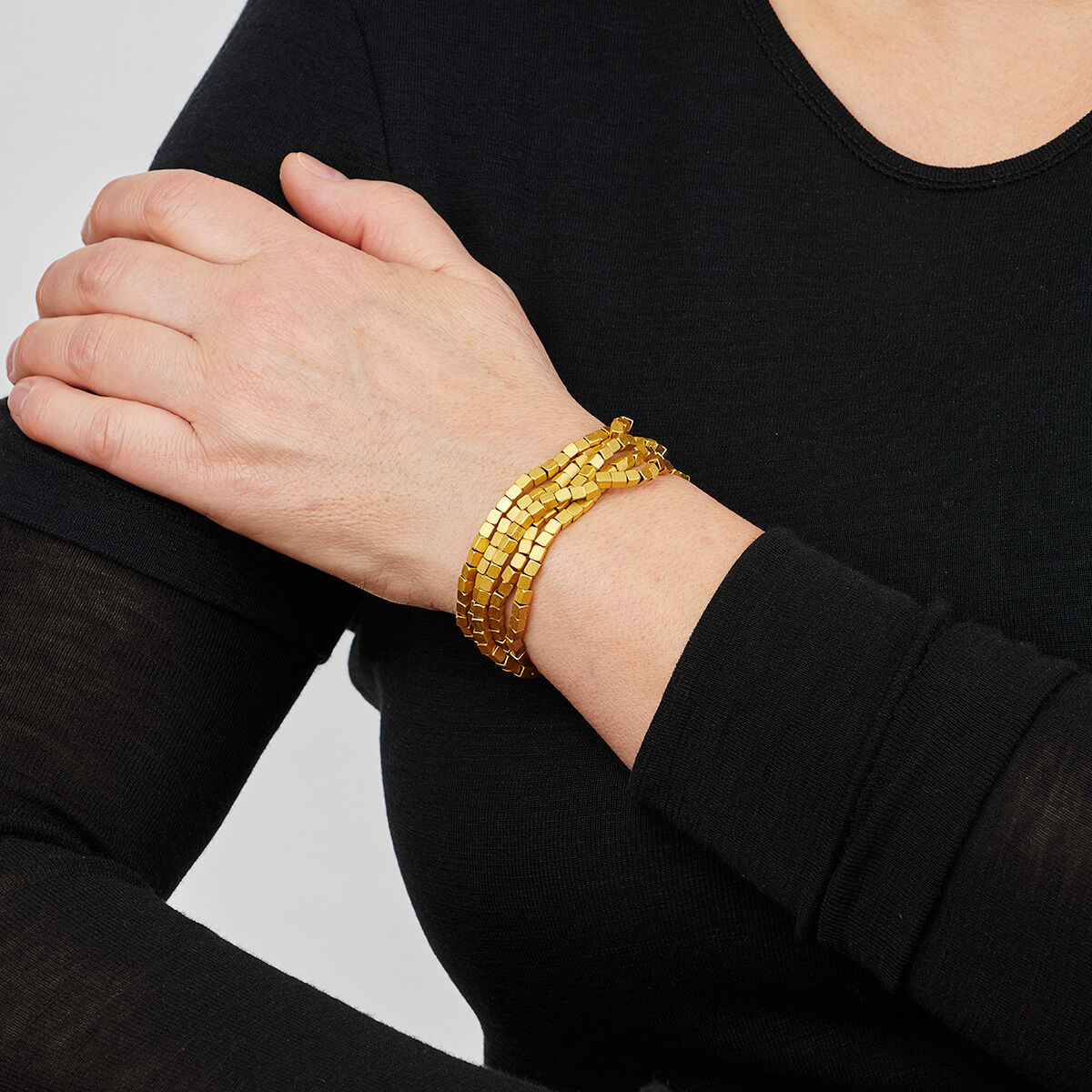 Nahaufnahme einer goldenen, würfelförmigen Kette, die das Handgelenk einer Person mit schwarzer Kleidung schmückt.