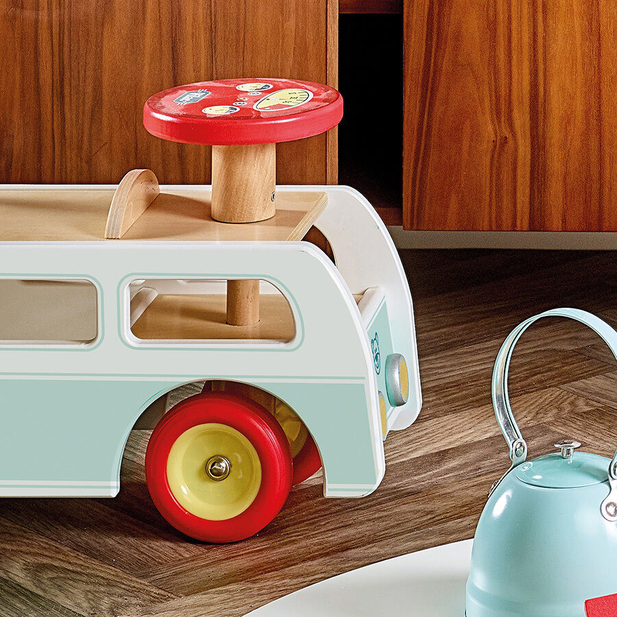 Ein Minibus aus Holz mit roten und gelben Rädern steht in einem Kinderzimmer. Der Minibus hat ein türkisfarbenes Dach und eine beige Karosserie. Im Hintergrund befindet sich ein Möbelstück aus Holz und ein türkisfarbener Wasserkocher.
