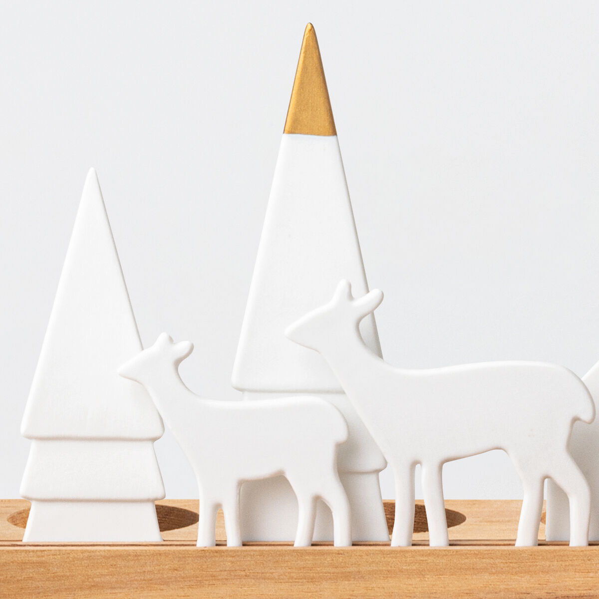 Das Bild zeigt ein stilisiertes Lichtobjekt namens »Wald«, bestehend aus weißen, geometrisch gestalteten Keramikfiguren von Bäumen, zwei Hirschen und einem Haus mit feinen Lichtdurchlässen, die auf einem schlichten Holzsockel angeordnet sind. Die Komposition vermittelt ein modernes, winterliches Waldmotiv und eignet sich als dekoratives Wohnaccessoire zur festlichen Jahreszeit.