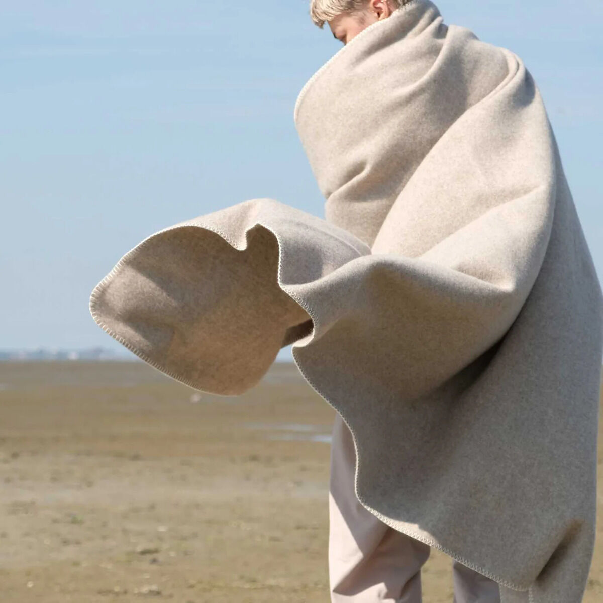 Eine Person ist in eine großzügige hellgraue Merinodecke mit weicher Textur gehüllt und steht auf einer freien, natürlichen Landschaft im Freien. Die Decke hat ein elegantes, minimalistisches Design und vermittelt einen Eindruck von Komfort und Geborgenheit.