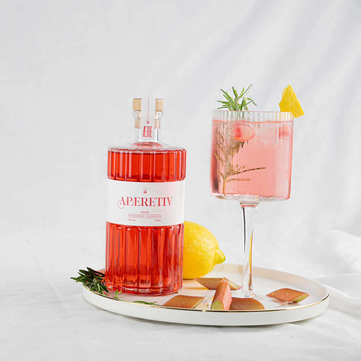 Eine stilvolle Flasche AERE Aperitif »Rosé« und ein dazugehöriges gefülltes Cocktailglas stehen auf einem Tablett. Dekoriert mit einer Zitronenscheibe, Rosmarin und einem Stück Grapefruit, vor einem weißen Hintergrund. Eine ganze Zitrone liegt ebenfalls auf dem Tablett.