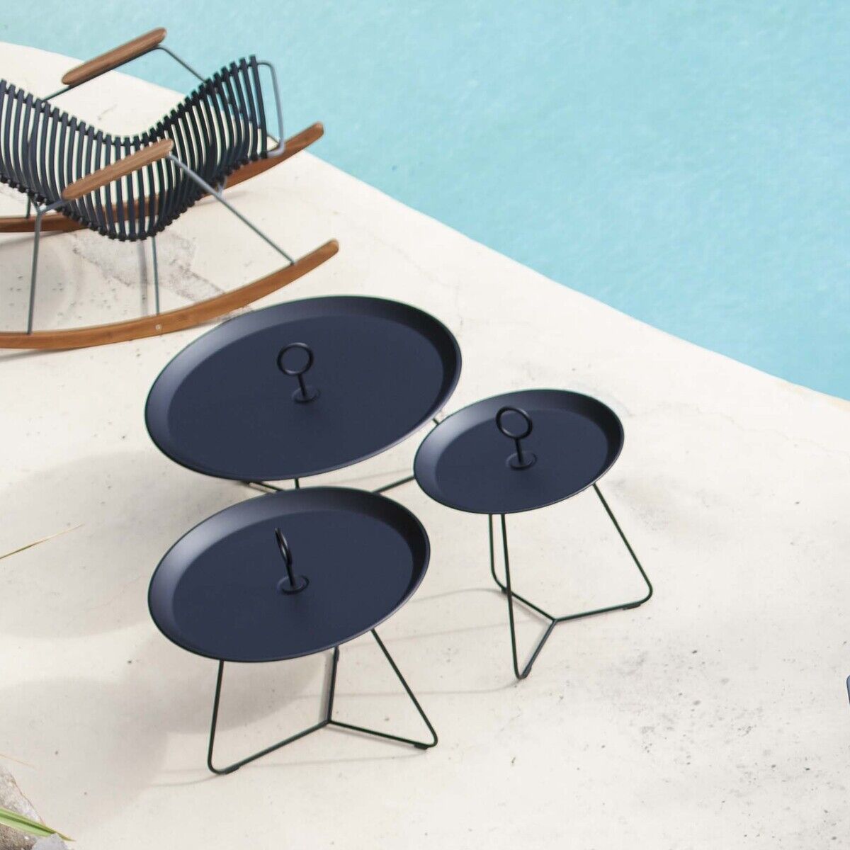 Eine Outdoor-Szene mit einem Pool im Hintergrund. Im Vordergrund steht ein runder Tisch mit Holzlatten und mehreren schwarz geflochtenen Stühlen. Zwei runde Tray Tables EYELET sind ebenfalls sichtbar, mit einem modernen und minimalistischen Design. Palmen und andere Pflanzen umgeben die Sitzmöbel.