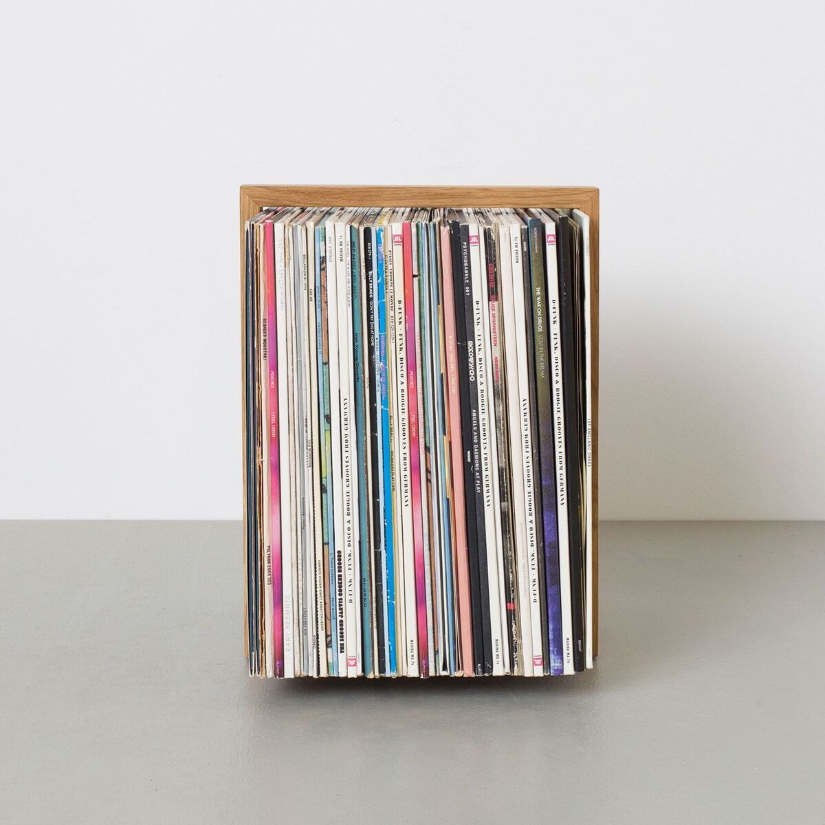 Ein stilvoller Schallplattenständer aus Holz, der eine Sammlung von Schallplatten ordentlich aufbewahrt und präsentiert. Der Ständer hat ein minimalistisches Design und steht auf einer grauen Fläche vor einer weißen Wand.