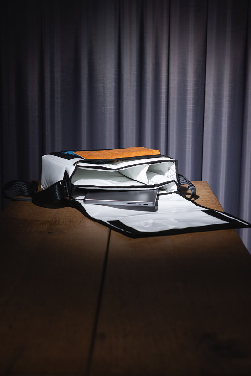 Eine moderne ZEIT-Tasche in Kastenform mit schwarzem Gurt. Die Tasche ist geöffnet und zeigt ein Laptop und ein Notizbuch. Die Tasche ist vor einem Vorhang auf einem Holztisch platziert.