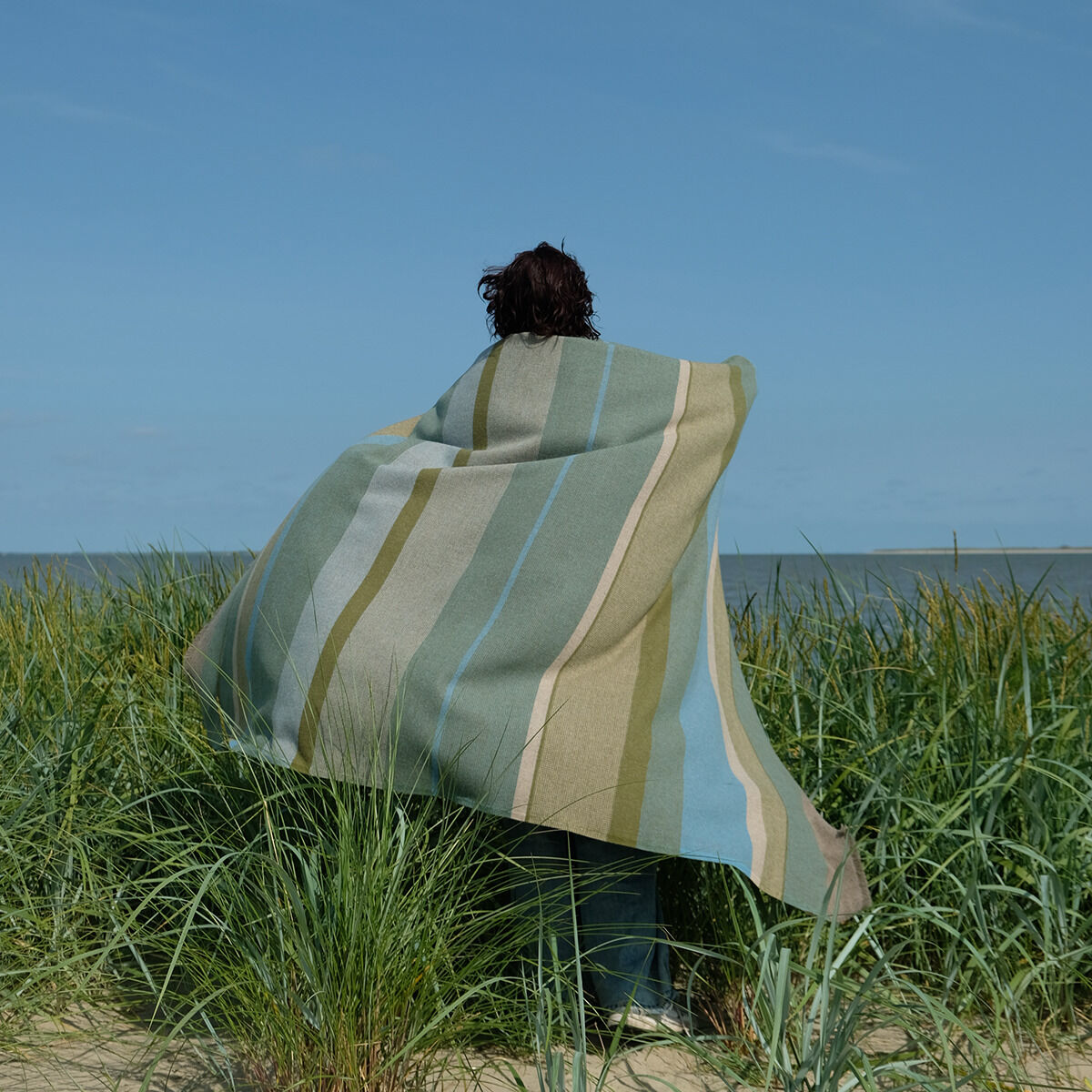 Eine Person steht am Strand und ist in eine gestreifte Merinowolldecke gehüllt. Die Decke zeigt natürliche Töne wie Blau, Grün und Beige. Im Hintergrund sind Gräser und das Meer zu sehen, was eine entspannte und natürliche Atmosphäre vermittelt.