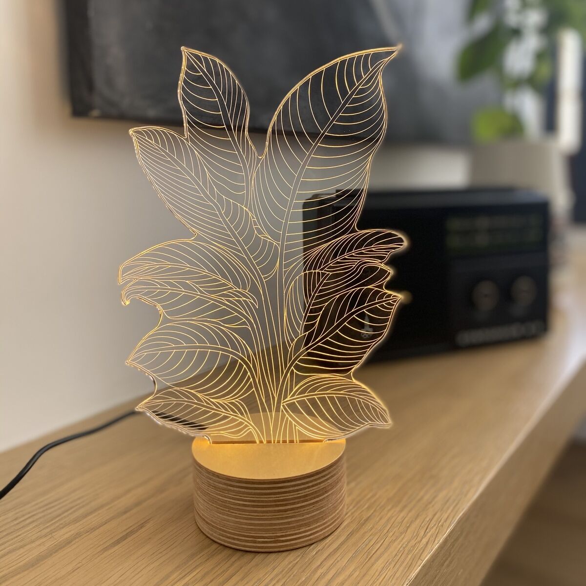 Eine 3D-Lampe mit Heliconia-Pflanzenmotiv auf einer Holzbasis, die auf einem hellen Holztisch steht. Im Hintergrund sind unscharfe Pflanzen und ein schwarzes technisches Gerät zu sehen.