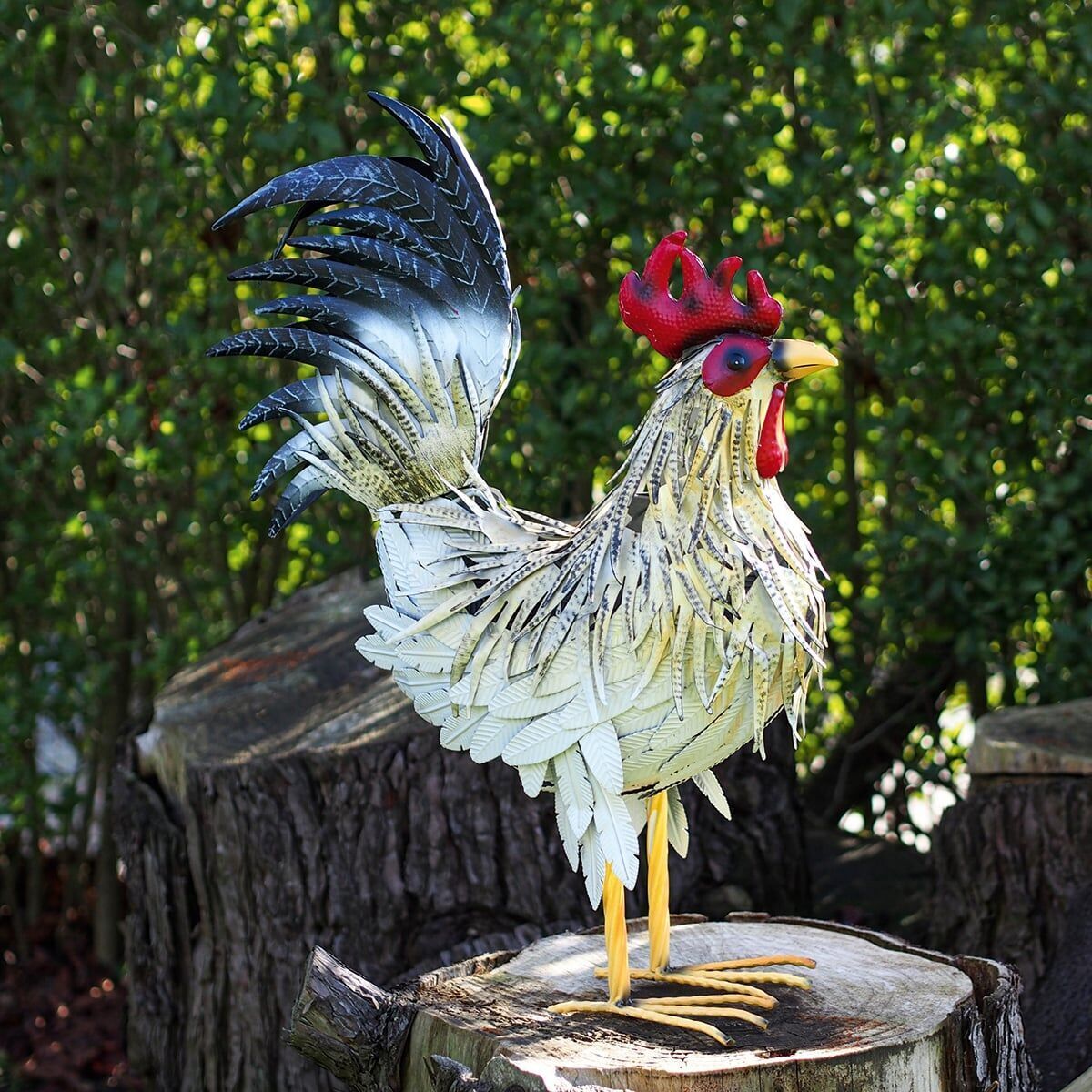 Dekorative Gartenfigur eines Hahns namens Fritz, aus Metall gefertigt, mit roten, gelben, grünen, schwarzen und weißen Farbakzenten, auf einem Baumstumpf vor einem Hintergrund aus grünen Büschen positioniert.