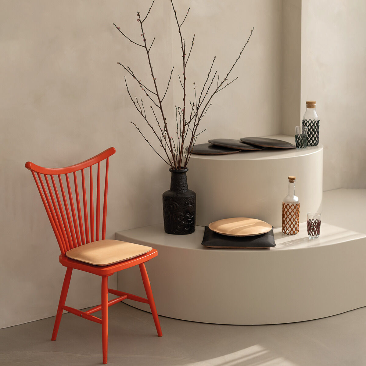 Stilvolles, helles Interieur mit einem roten Holzstuhl, auf dessen Sitz ein flaches, quadratisches beigefarbenes Polster liegt. Daneben steht ein rundes, zweistufiges cremefarbenes Podest mit schwarzen und naturfarbenen Tellern, Glasflaschen und einer dunklen Vase mit kahlen Zweigen.