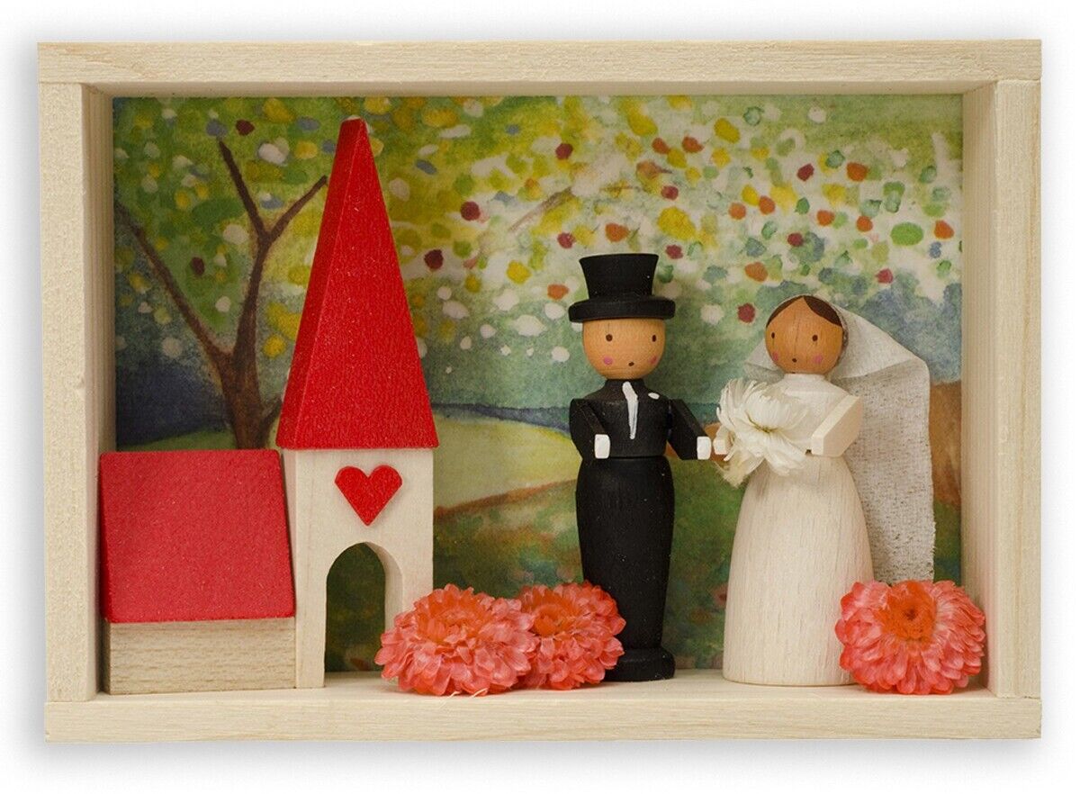 Glückwunschkästchen »Brautpaar«. Eine hölzerne Miniaturdarstellung eines Brautpaars. Im Hintergrund eine stilisierte Kirche mit rotem Dach und Herz. Weiße Blumen und grüne Landschaft im Hintergrund. Ideal als Hochzeits- oder Verlobungsgeschenk.
