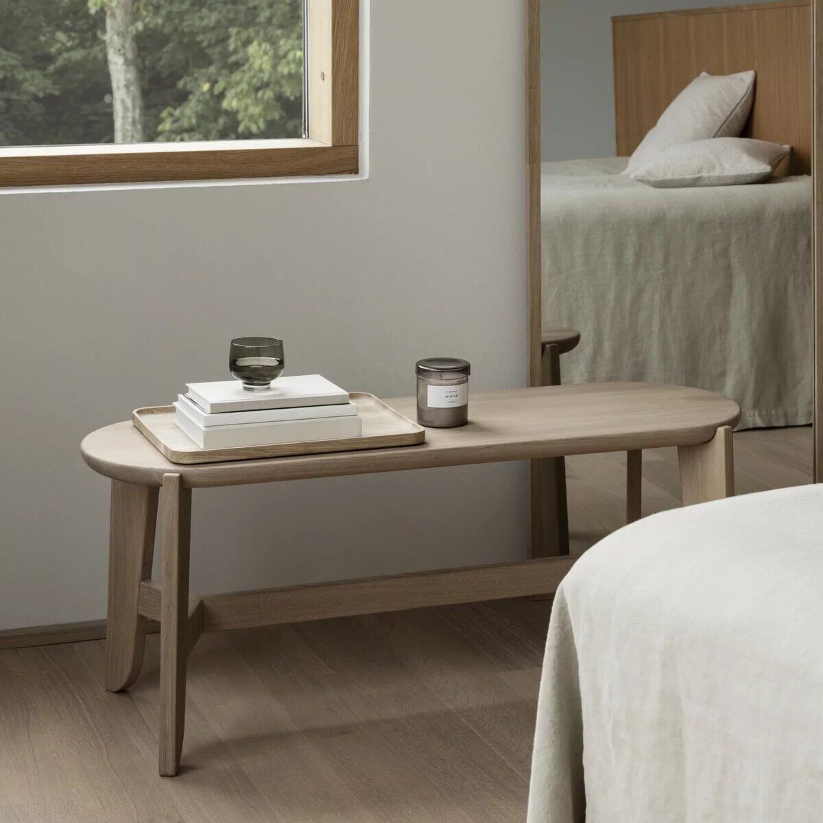Holz-Sitzbank im skandinavischen Stil steht in einem minimalistisch eingerichteten Schlafzimmer neben einem Bett, mit Dekoration wie Büchern und einer Kerze; natürlicher Lichteinfall durch das Fenster, ruhige und helle Farbgestaltung.