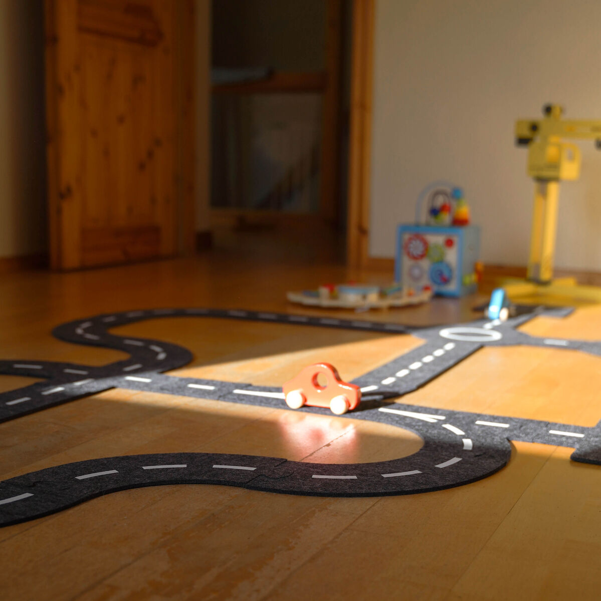 Filz-Spielstraße auf einem Holzboden, mit zwei kleinen Holzfahrzeugen. Die Spielstraße bildet verschiedene Kurven und Straßenverläufe. Im Hintergrund sind weitere Kinderspielzeuge und ein weicher Lichteinfall zu sehen.