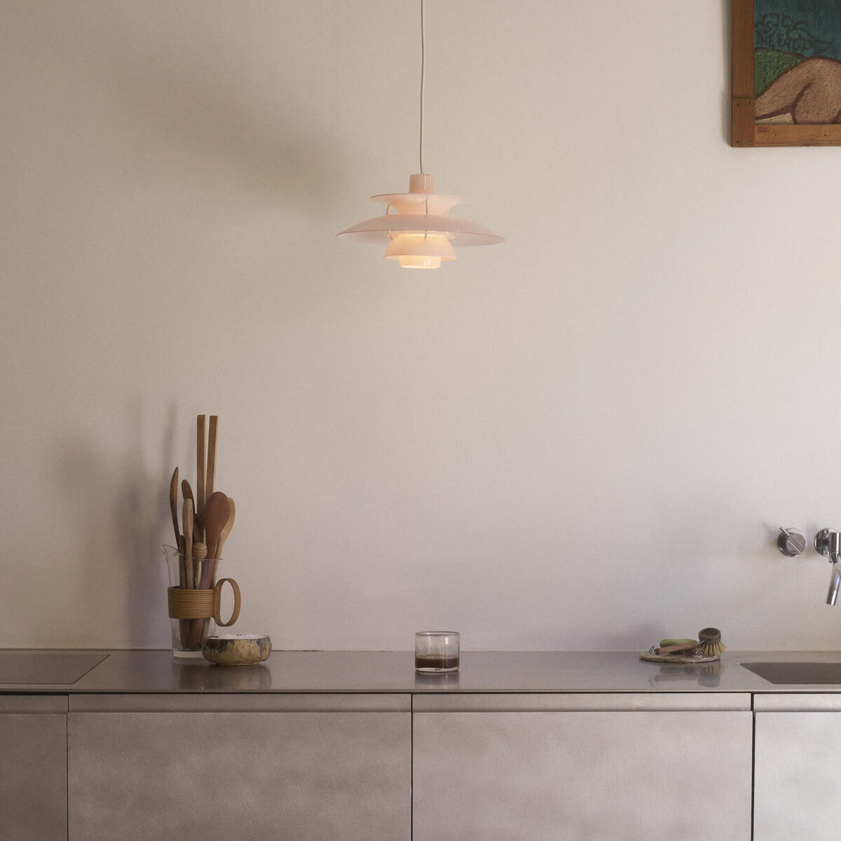 Hängeleuchte »PH 5 320« von Louis Poulsen in dezentem Opal-Rosa hängt über einer modernen Edelstahl-Anrichte. Harmonisches Licht strahlt nach unten und diffus in den Raum. Zubehör wie Holzlöffel im Becher und kleine Schalen ergänzen das stilvolle Ambiente.