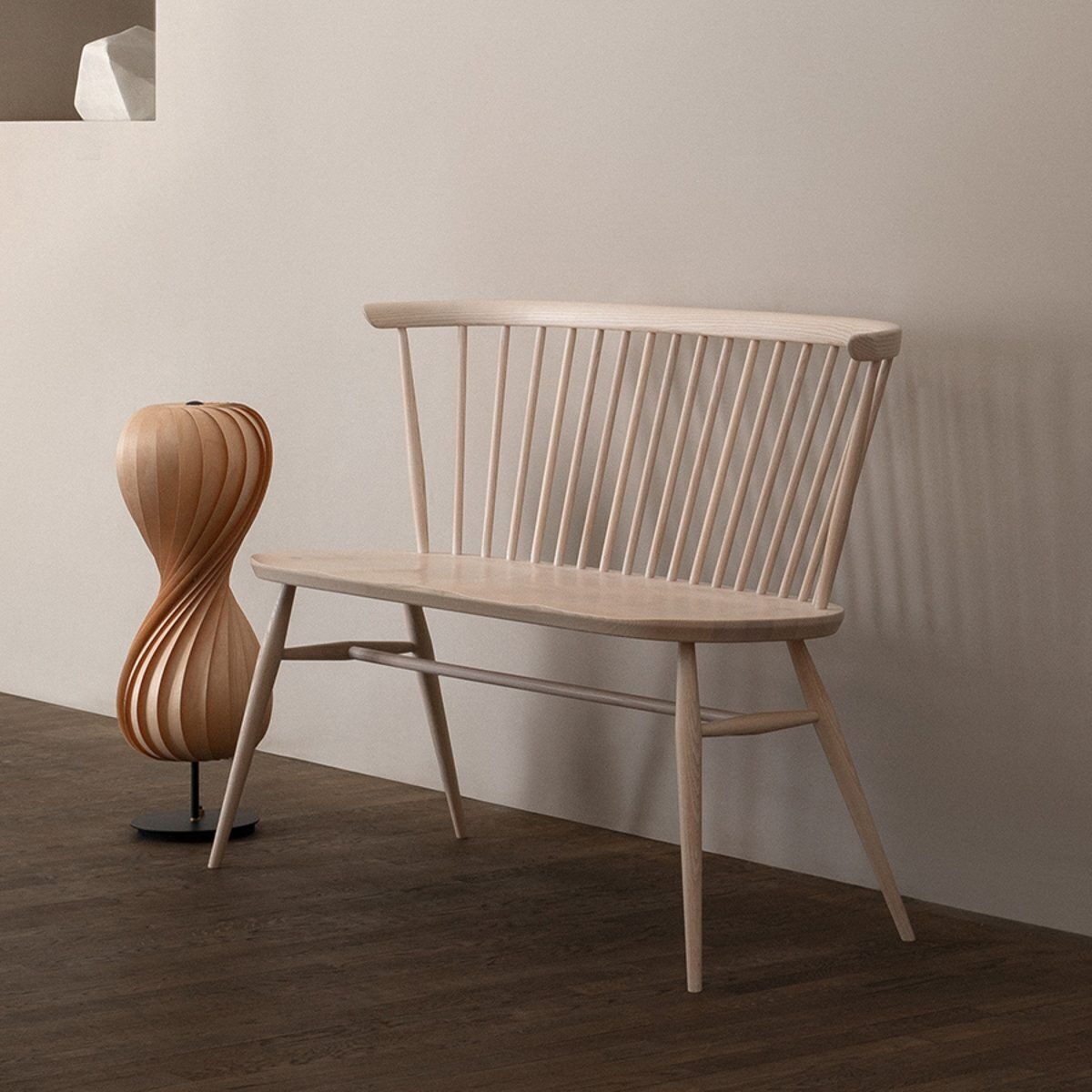 Eine stilvolle Holzbank aus hellem Holz, bekannt als die ZEIT-Sonderedition »Loveseat« von Ercol. Sie steht vor einer beige Wand und einem dunkelbraunen Holzboden, begleitet von einer Designer-Stehlampe aus Holz.