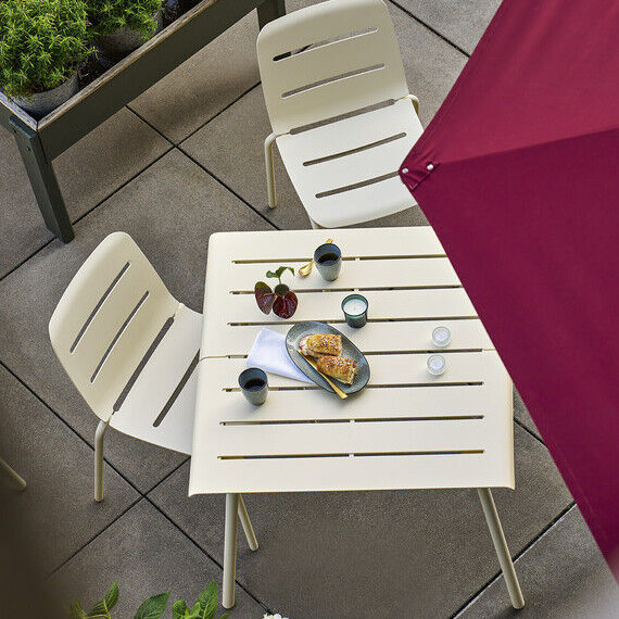 Ein moderner weißer Tisch mit vier passenden Stühlen steht auf einer Terrasse. Auf dem Tisch befinden sich Getränke und Snacks. Ein großer roter Sonnenschirm spendet Schatten. Im Hintergrund sind Pflanzen zu sehen.