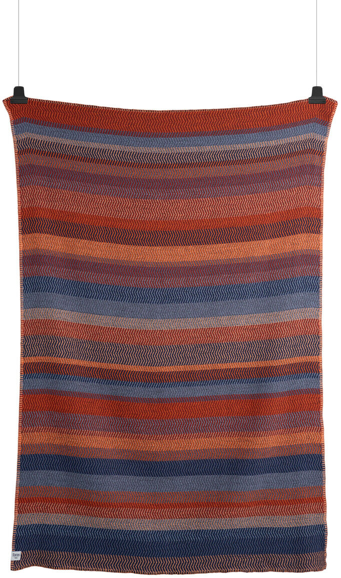 Gestreifte Lammwolldecke mit roten, blauen, orangefarbenen und braunen Streifen, die an einer Wand hängt.