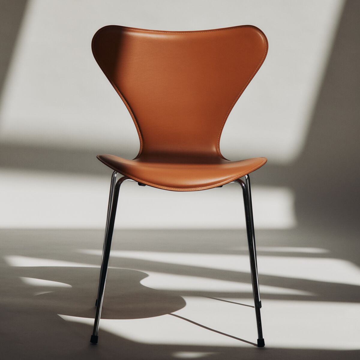 Der Stuhl »Series 7™ Tailored« von Fritz Hansen steht zentral in einem von Licht durchfluteten Raum. Die Sitzfläche und Rückenlehne sind in sattem Sattelbraun gepolstert, das elegante Untergestell besteht aus verchromtem Metall mit schlanken schwarzen Füßen. Scharfe Licht- und Schattenkanten am Boden betonen das ikonische Design.