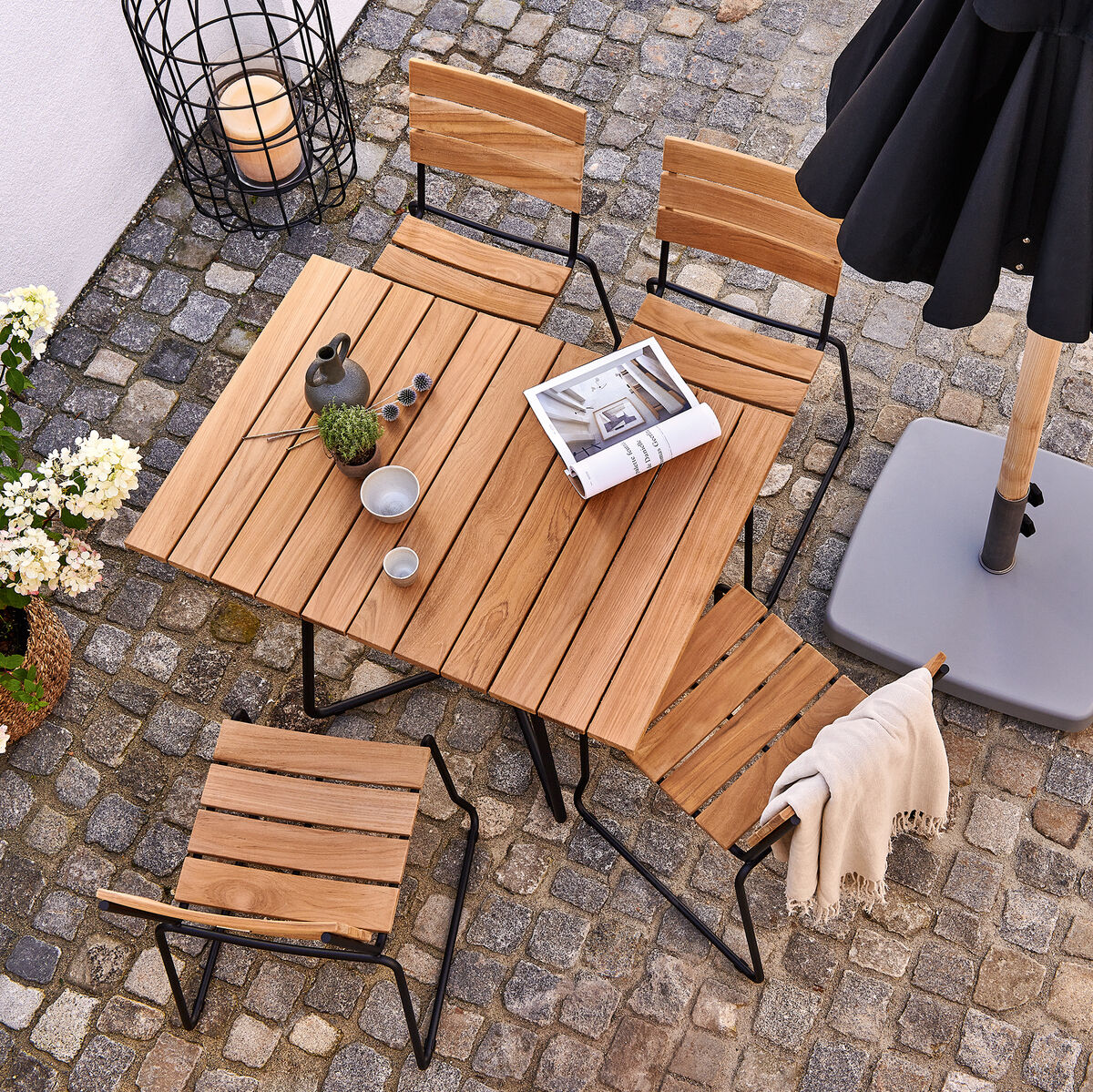 Vier Teakholzstühle »BALCONY« mit Metallgestell und ein passender quadratischer Tisch stehen auf einem gepflasterten Boden. Eine Laterne, eine Pflanze, Kerzen und ein Buch dienen als Dekoration. Ein schwarzer Sonnenschirm spendet Schatten.