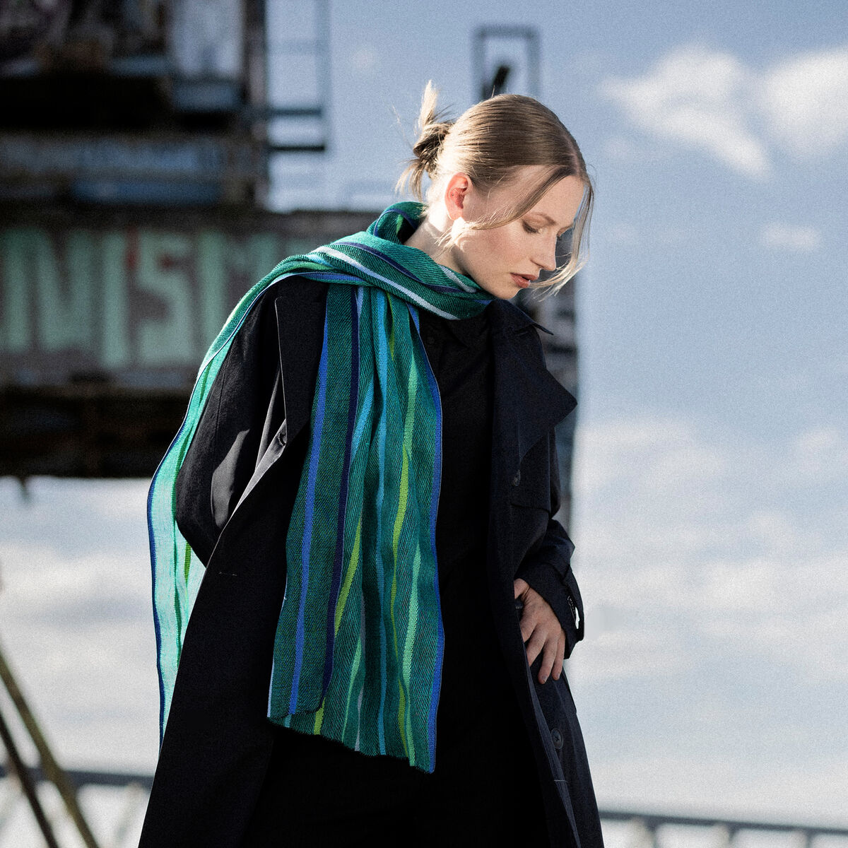 Eine Frau trägt einen gestreiften Leinenschal von Ulrike Isensee in Grün und Blau über einem dunklen Mantel. Der Schal wirkt leicht und elegant vor einem industriellen Hintergrund mit blauem Himmel und sanften Wolken.