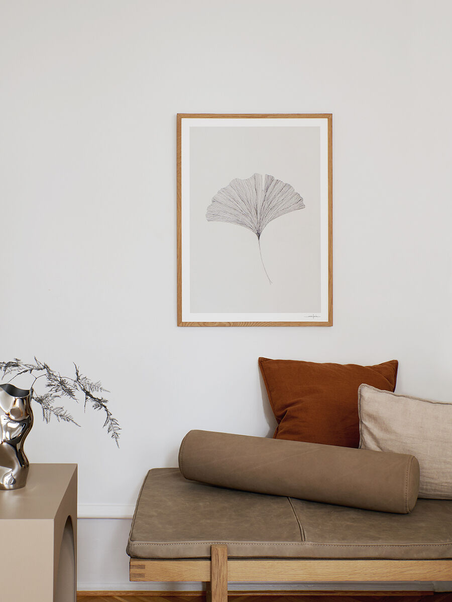 Gerahmtes Bild eines Ginkgoblattes an einer weißen Wand. Darunter ein Sitzbereich mit beigen und braunen Kissen und einer grauen Rolle. Ein silbernes Dekogefäß steht auf einem Tisch.