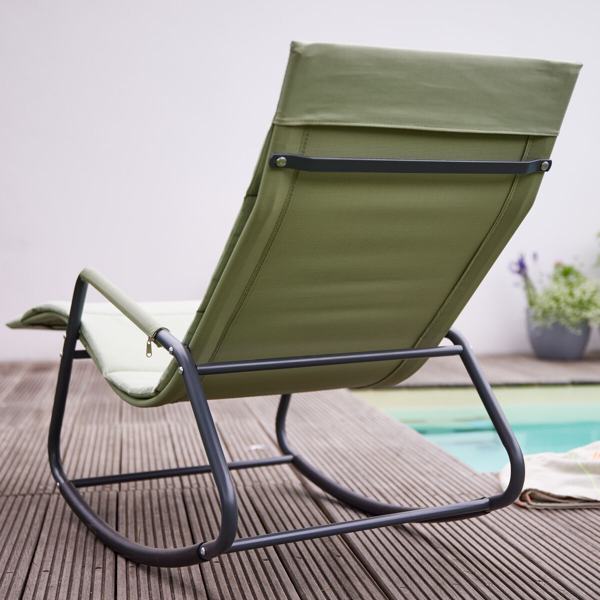 Eine moderne olivgrüne Swingliege »Aliano« mit schwarzem Gestell steht auf einer Holzterrasse neben einem Pool. Im Hintergrund sind Pflanzen in Töpfen zu sehen. Auf der Liege liegt ein Handtuch und eine Sonnenbrille.