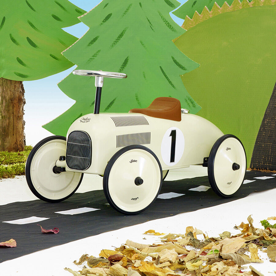 Ein Spielzeug Vintage-Auto aus Metall, in weißer Farbe mit schwarzen Rädern und der Nummer 1 an den Seiten. Es steht auf einer Straße und wird von grün gefärbten Büschen und einem Baum im Hintergrund umgeben. Das Auto hat einen braunen Sitz und ist für Kinder geeignet.
