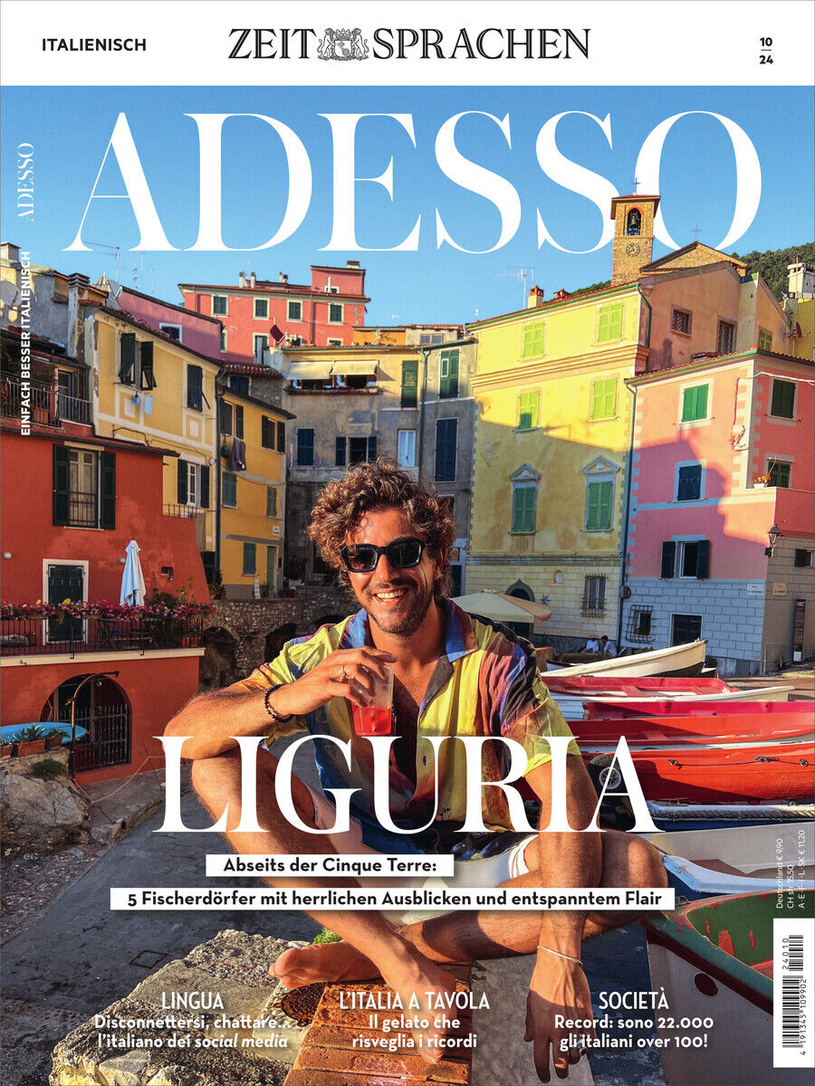 Titelbild des Adesso Magazins 10/2024 zeigt einen Mann in sommerlicher Kleidung, der vor bunten, mediterranen Gebäuden in Ligurien, Italien sitzt. Im Hintergrund sind Fischerboote und die Abendsonne zu sehen.