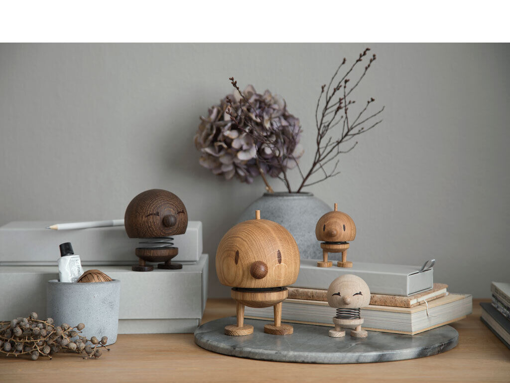 Ein Arrangement von Hoptimist Bumble Figuren aus Holz in verschiedenen Größen stehen auf einem Tisch vor einem grauen Hintergrund. Eine große Figur aus Eichenholz ist in der Mitte platziert, umgeben von weiteren Figuren in unterschiedlichen Holztönen. Im Hintergrund befinden sich getrocknete Blumen und Bücherstapel, die eine gemütliche und stilvolle Wohnatmosphäre schaffen.
