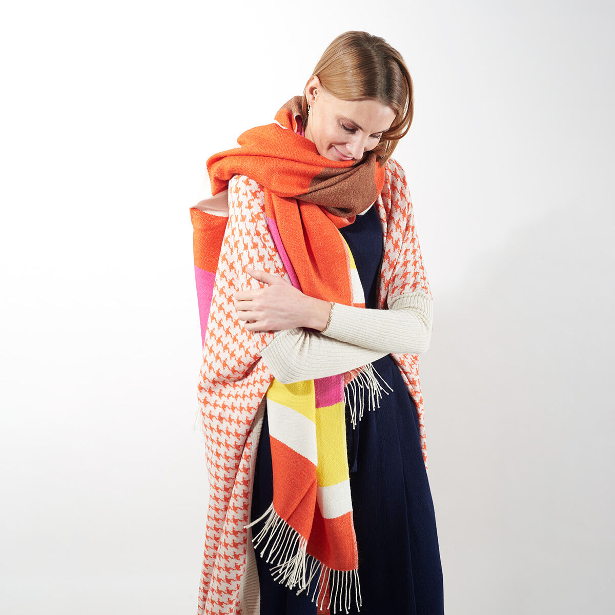 Eine Frau trägt das ZEIT-Jubiläumsedition Cape »Surya« mit farbenfrohen Mustern in Orange, Rosa, Braun, Weiß und Gelb, kombiniert mit einem dunkelblauen Kleid. Das Cape wirkt modisch und komfortabel.