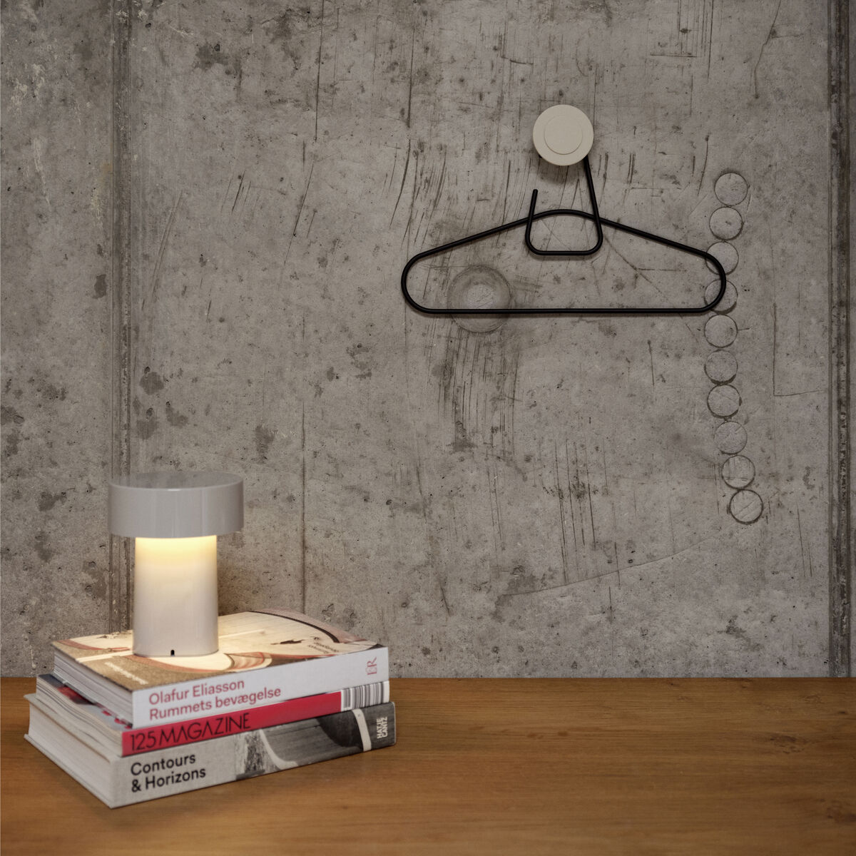 Eine tragbare, moderne Lampe mit zylindrischem Lampenschirm steht auf einem Stapel Bücher auf einer Holzoberfläche. Im Hintergrund ist eine graue Betonwand mit einer minimalistischen Garderobe und einigen Metallhaken zu sehen. Die Lampe strahlt ein warmes, angenehmes Licht aus und sorgt für eine gemütliche Atmosphäre.