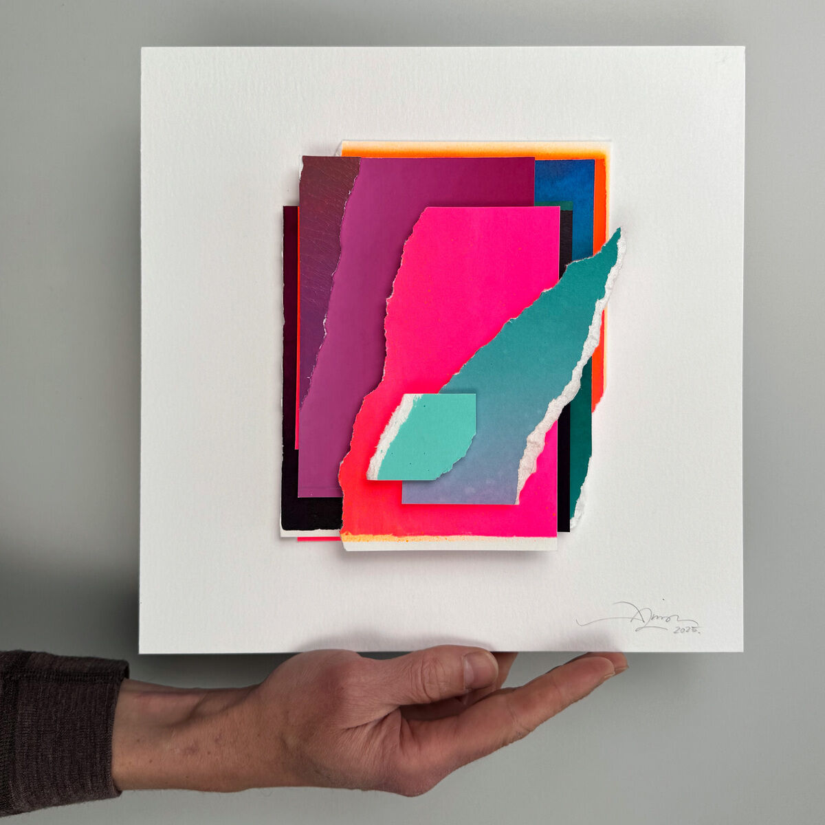 Quadratische Collage von Daniel Lisson mit farbigen, teils gerissenen Papierstücken in Pink, Magenta, Cyan, Türkis, Orange und Weiß auf weißem Grund. Die Collage ist dreidimensional gerahmt, eine Hand hält das Bild. Rechts unten Signatur des Künstlers.