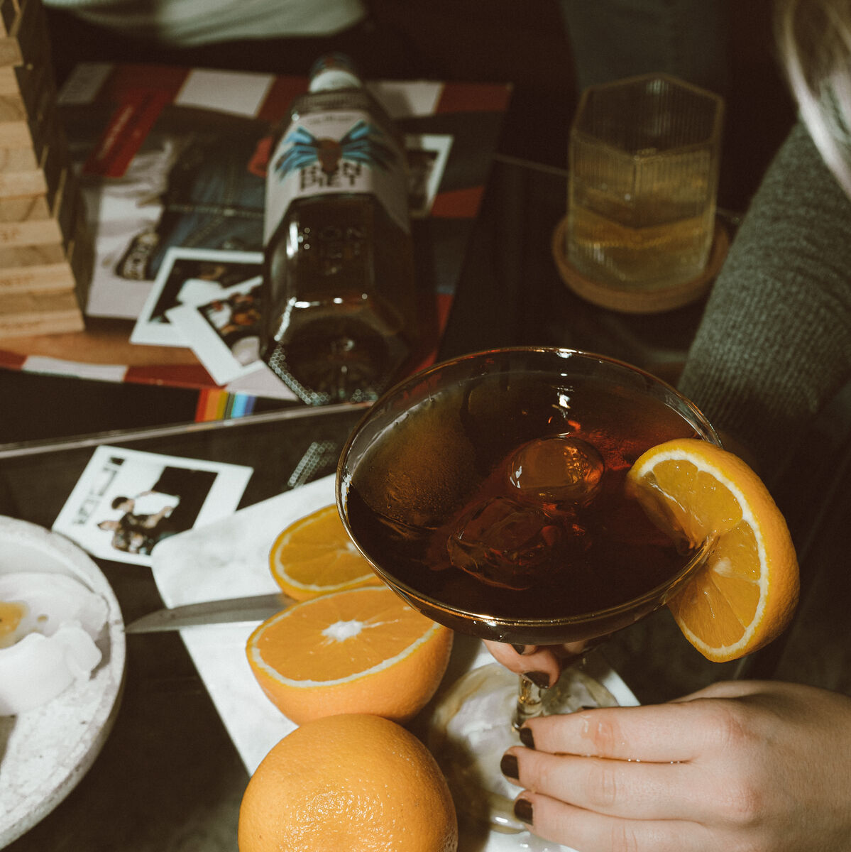 Eine Flasche Ron Piet Rum, 10 Jahre gereift, steht auf einem Tisch mit Cocktails und Orangenscheiben. Ein Glas mit einem Cocktail und Orangenscheiben wird von einer Hand gehalten. Polaroidfotos und verschiedene Gegenstände befinden sich ebenfalls auf dem Tisch.