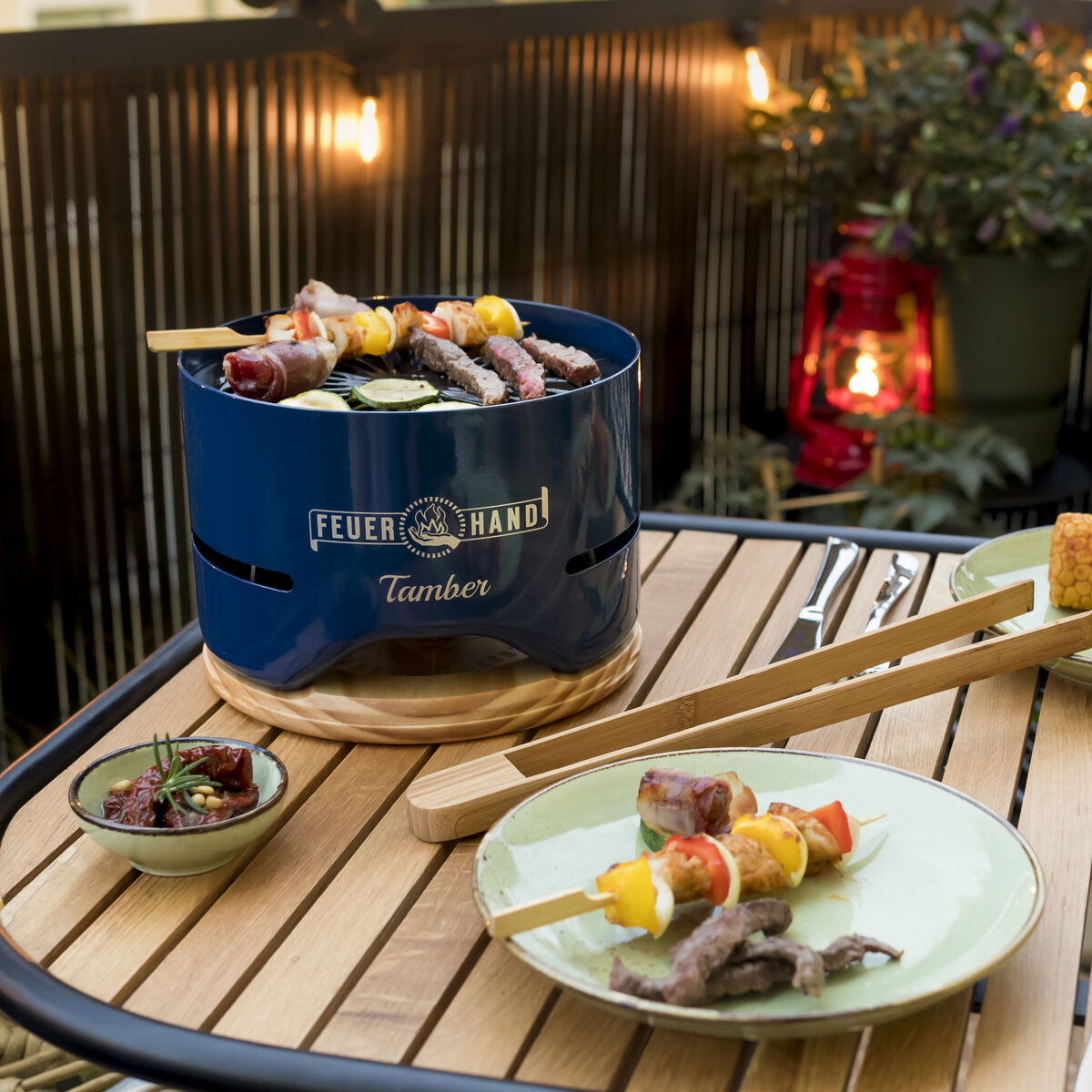 Ein Tischgrill namens Tamber steht auf einem Tisch auf einem Balkon, umgeben von verschiedenen Grillspießen, Maiskolben, zwei Gläsern Rotwein und einem Brotkorb. Im Hintergrund ist eine gemütliche Balkonbeleuchtung zu sehen.