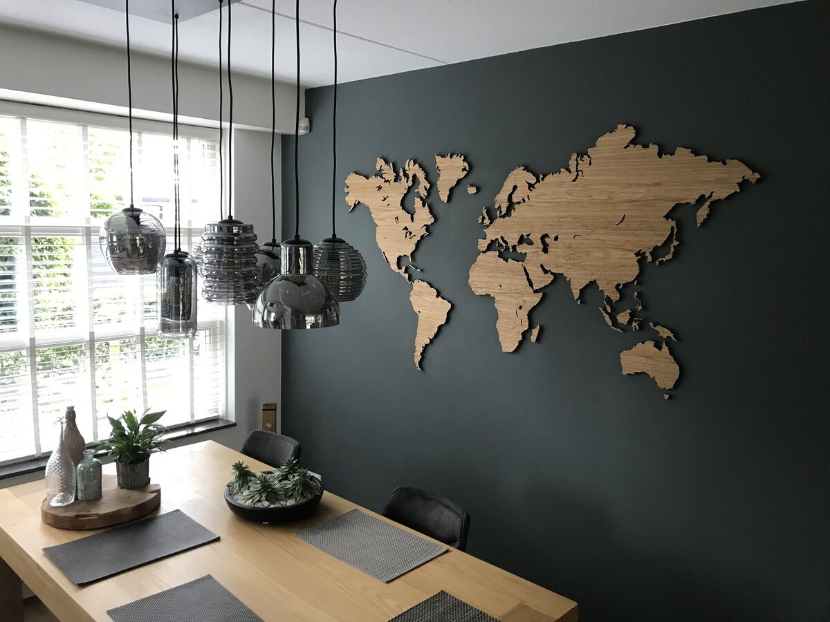 An einer dunkelgrauen Wand hängt eine detaillierte Weltkarte aus Eichenholz. Auf dem darunterliegenden Holztisch befinden sich Dekorationen wie Pflanzen und Glasgefäße. Mehrere moderne Hängelampen beleuchten den minimalistischen Essbereich.