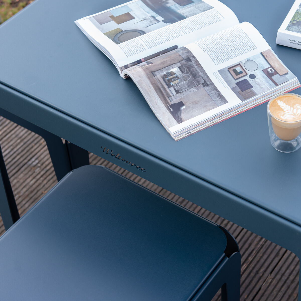 Ein quadratischer Gartentisch im modernen Design, auf dem eine Tasse Kaffee und zwei offene Magazine liegen. Der Tisch besteht aus wetterfestem Metall, geeignet für den Außenbereich.