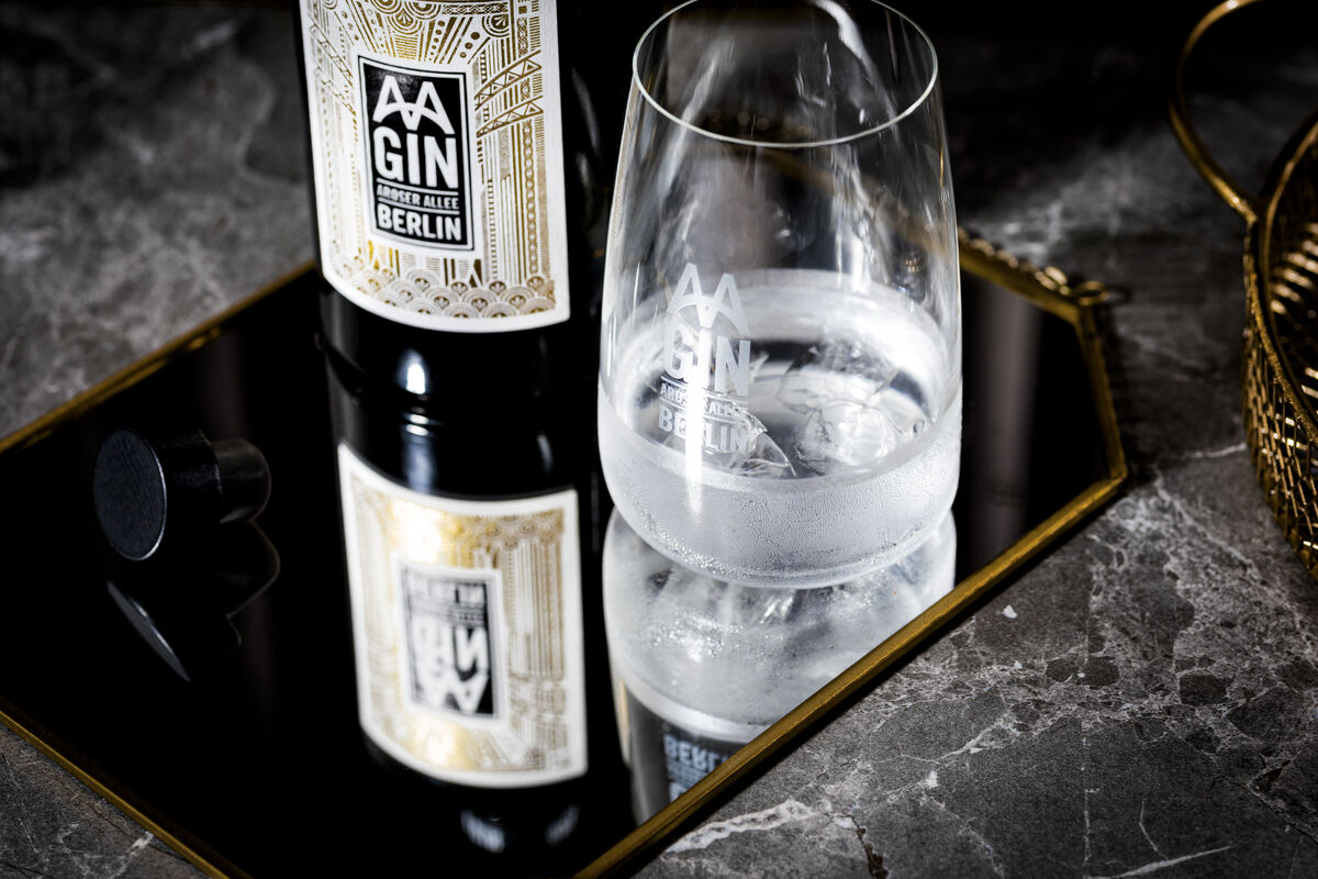 Eine Flasche »AAGIN« Dry Gin und ein Glas im Set präsentiert auf einer reflektierenden Oberfläche mit eleganter, goldener Umrandung, ideal für luxuriöse Anlässe.