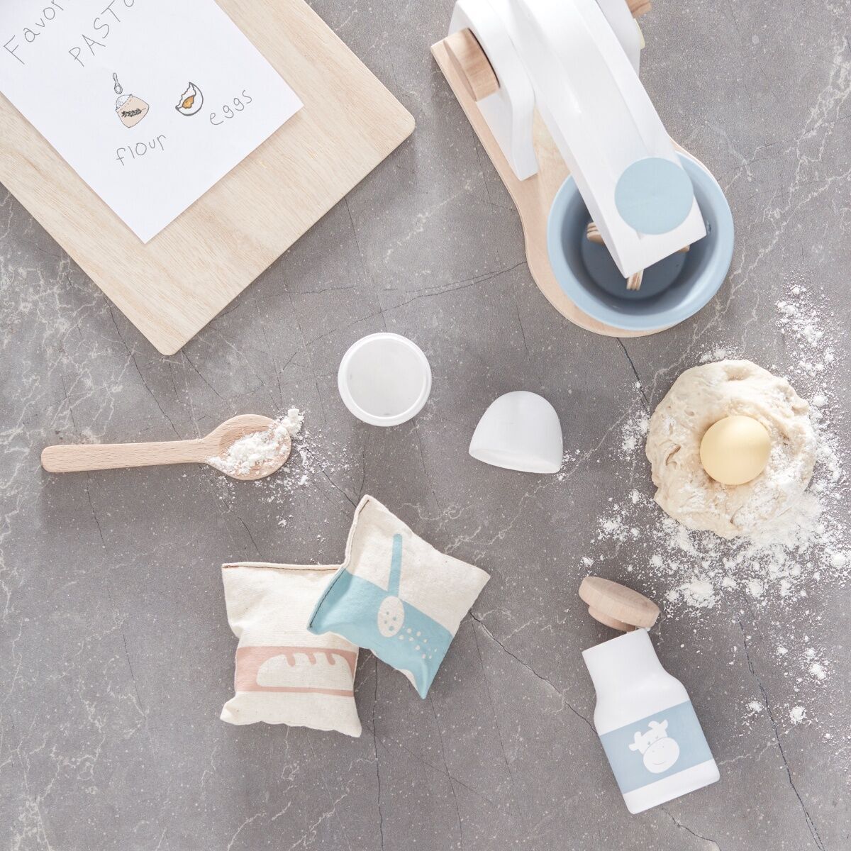 Ein Holzmixer-Set samt Zubehör liegt auf einer grauen Oberfläche. Das Set enthält unter anderem einen Holzmixer, eine Schüssel, ein Ei, Mehlbeutel und eine Milchflasche. Neben dem Mixer liegt ein Brett mit einer Rezeptkarte und einem Messlöffel.