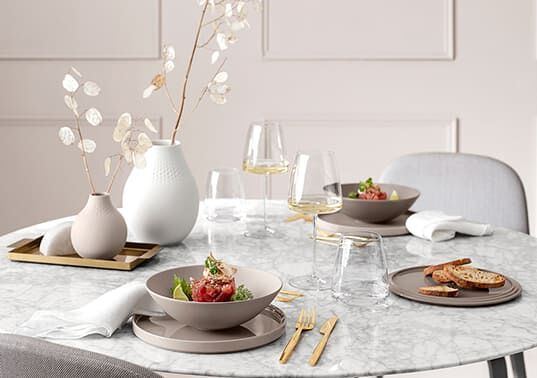 Eleganter gedeckter Tisch mit grauen Tellern, goldfarbenem Besteck, Weingläsern und weißen Vasen mit Zweigen