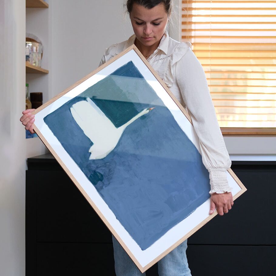 Eine Frau hält ein gerahmtes Poster mit einer stilisierten, minimalistischen Illustration eines weißen Schwans auf blauem Hintergrund von Jörgen Hansson. Die Szene spielt in einem modern eingerichteten Raum mit hellen Farben und Fenster im Hintergrund.