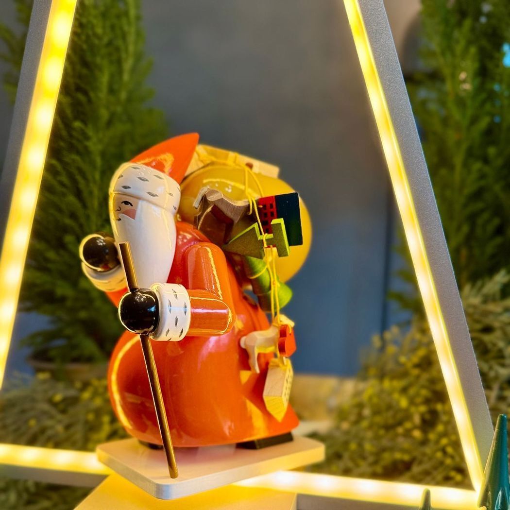 Detailreiche Holz-Figur »Großer Weihnachtsmann mit Spielzeug« der Marke Wendt & Kühn. Der Weihnachtsmann trägt rotes Gewand und Mütze, hält einen goldenen Stab und hat einen Sack voller bunter Geschenke. Umgeben von warmen Lichtern und grüner Tanne auf einer leuchtenden Basis.
