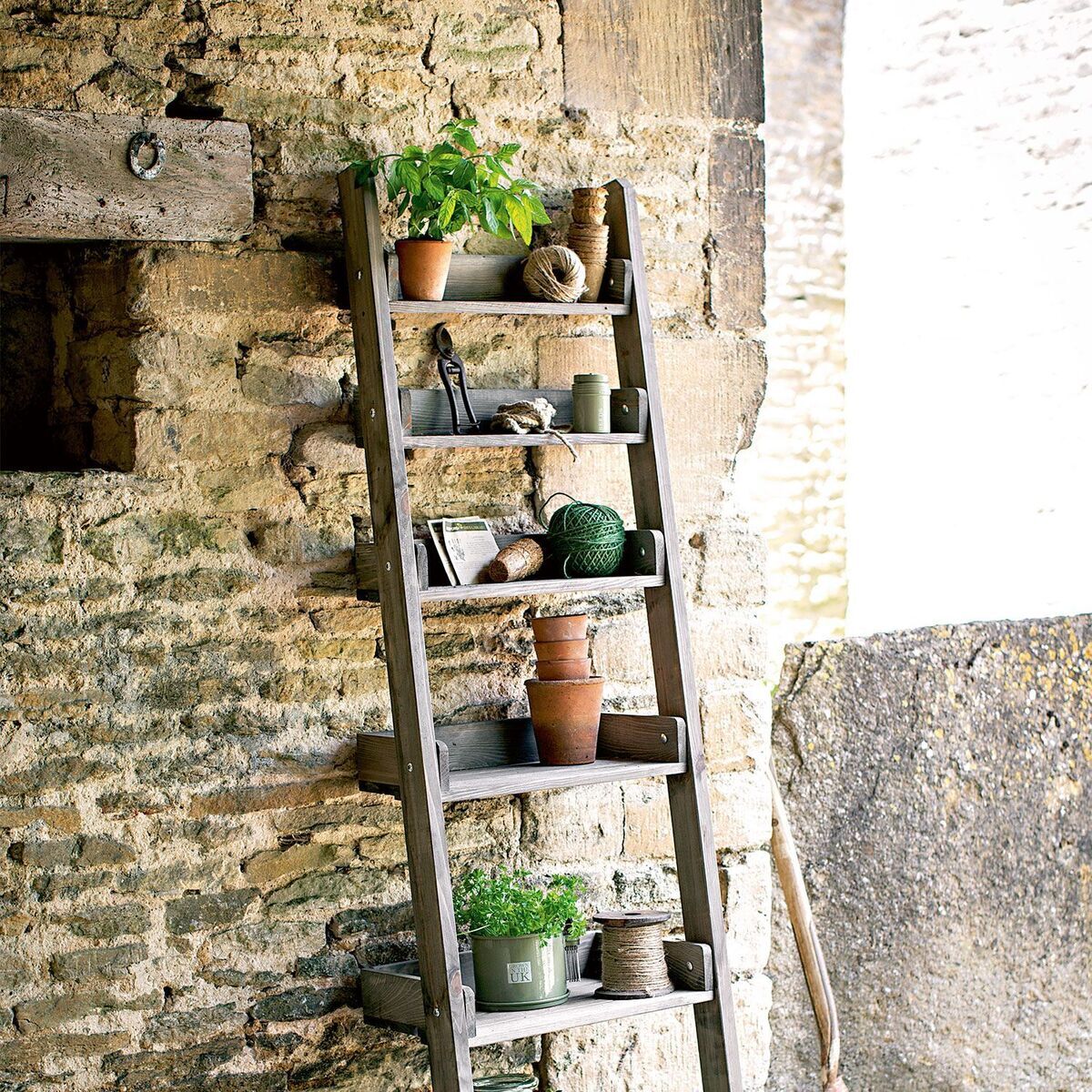 Ein rustikales Holzregal im Landhausstil, das zum Anlehnen an eine Steinmauer konzipiert ist. Es enthält verschiedene Blumentöpfe und Kräuter auf mehreren Ebenen.