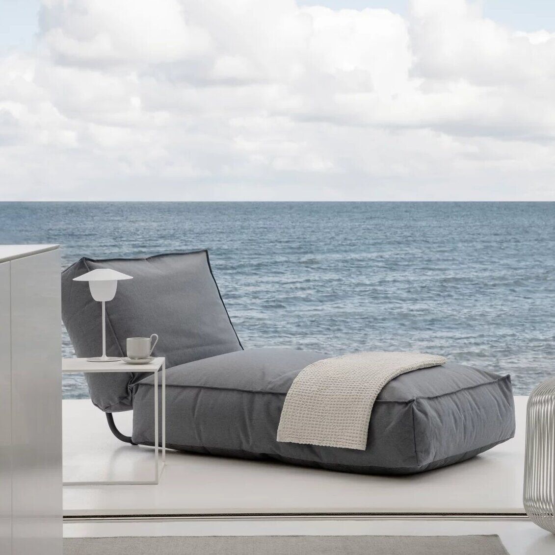 Ein Outdoor-Bett 'STAY' mit den Maßen 80 x 190 cm steht auf einer Terrasse mit Meerblick. Das Bett ist mit grauem Stoff bezogen und mit einer beigen Decke und einem weißen Kissen ausgestattet. Neben dem Bett steht ein kleiner weißer Beistelltisch mit einer Lampe und einer Tasse. Der Himmel ist klar mit einigen Wolken sichtbar.