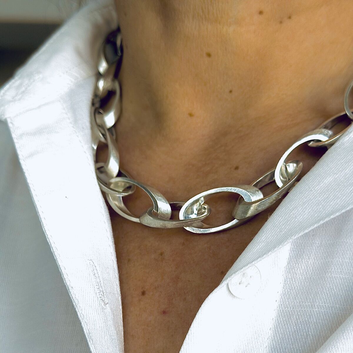 Nahaufnahme eines silbernen Colliers namens "Jackie" um den Hals einer Person, getragen über einem weißen Hemd. Die Kette besteht aus ovalen Gliedern, die elegant und modern wirken.