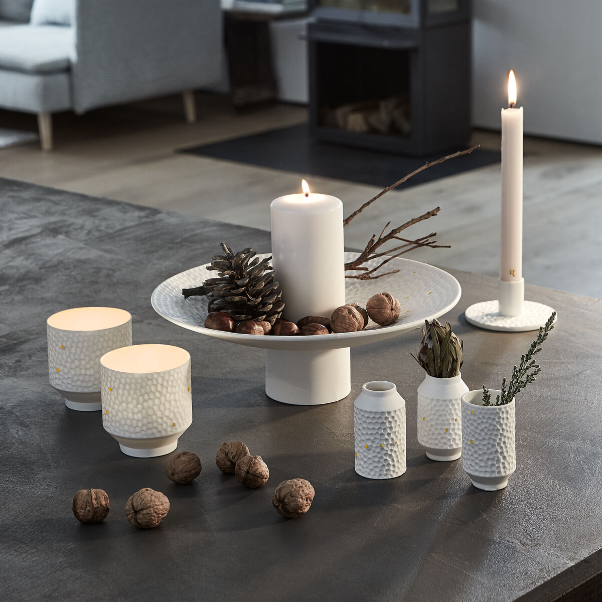 Im Vordergrund steht die weiße Deko-Schale »Sternenbild« von Räder, dekoriert mit Zapfen, Nüssen und Kerzen, auf einem dunklen Tisch. Im Hintergrund modernes Wohnzimmer mit Sofa, Kaminofen und minimalistisch eingerichteter Umgebung.